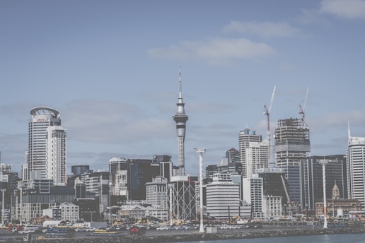 A modern city skyline featuring a mix of high-rise buildings, including a prominent tower with a slender structure. Construction cranes are visible, indicating ongoing development. The sky is partly cloudy, and the waterfront is lined with various structures.