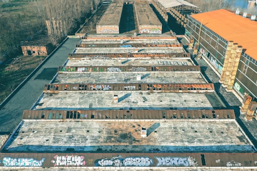 An aerial view of an industrial area featuring several large, rectangular buildings with flat roofs, covered in graffiti. The buildings are surrounded by bare trees and patches of vegetation. There are industrial chimneys visible on the right side.