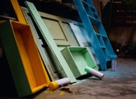 A collection of colorful wooden structures and spray paint cans are scattered on a concrete surface. The structures include a bright yellow open box, a light green door, and a large blue object resembling a ladder or shelf. The scene suggests an artistic or DIY environment.