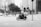A black and white photo captures an urban scene with a motorcyclist carrying a large load of packages. The background features modern buildings, pedestrians walking on the sidewalk, and cars on the street. The trees and city infrastructure add to the bustling city atmosphere.