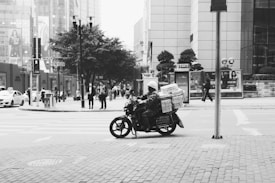 A black and white photo captures an urban scene with a motorcyclist carrying a large load of packages. The background features modern buildings, pedestrians walking on the sidewalk, and cars on the street. The trees and city infrastructure add to the bustling city atmosphere.