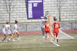 A lacrosse match with players actively engaged near the goal. One player in a white uniform with the number 33 is guarding the goal, while two players in red uniforms with numbers 20 and 30 are playing offensively. Three players in white are approaching with lacrosse sticks. A referee is observing nearby. A banner with championship years is visible in the background against a stone wall.