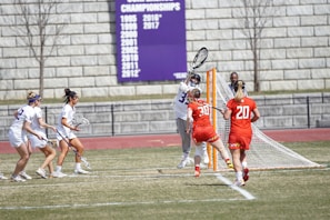 A lacrosse match with players actively engaged near the goal. One player in a white uniform with the number 33 is guarding the goal, while two players in red uniforms with numbers 20 and 30 are playing offensively. Three players in white are approaching with lacrosse sticks. A referee is observing nearby. A banner with championship years is visible in the background against a stone wall.