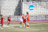A group of female lacrosse players is on a grassy field near a brick wall with an NCAA sign. They are wearing red and white uniforms, with some players holding lacrosse sticks. The scene captures movement and teamwork, possibly during a game.