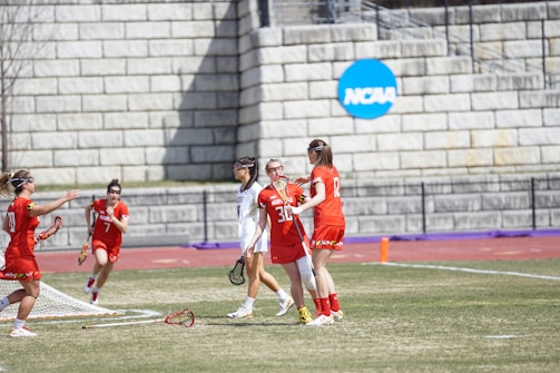 A group of female lacrosse players is on a grassy field near a brick wall with an NCAA sign. They are wearing red and white uniforms, with some players holding lacrosse sticks. The scene captures movement and teamwork, possibly during a game.