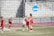 A group of female lacrosse players is on a grassy field near a brick wall with an NCAA sign. They are wearing red and white uniforms, with some players holding lacrosse sticks. The scene captures movement and teamwork, possibly during a game.