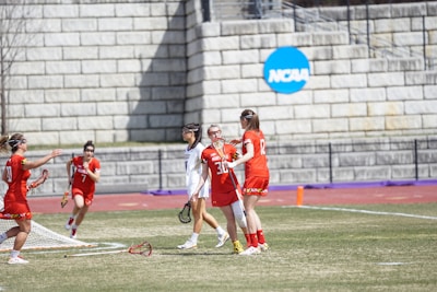 A group of female lacrosse players is on a grassy field near a brick wall with an NCAA sign. They are wearing red and white uniforms, with some players holding lacrosse sticks. The scene captures movement and teamwork, possibly during a game.