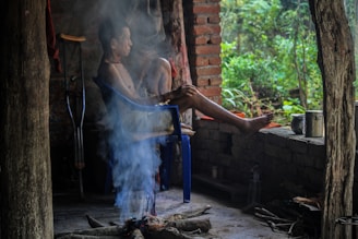 A man sits on a blue plastic chair inside a rustic, open-walled structure with brick and wooden elements. He appears to be relaxing or meditating with legs crossed and crutches by his side. There is a fire pit in front of him with smoke rising, and a view of lush greenery outside.