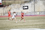 Players are engaged in a competitive lacrosse game on a grassy outdoor field. One player in a red uniform is running with the ball, pursued closely by an opponent in a white uniform. A scoreboard and stone bleachers are visible in the background.