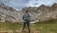 A person wearing a backpack stands on a grassy landscape, looking at snow-dappled mountain peaks under an overcast sky, conveying a sense of adventure and exploration.