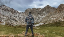 A person wearing a backpack stands on a grassy landscape, looking at snow-dappled mountain peaks under an overcast sky, conveying a sense of adventure and exploration.