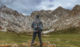 A person wearing a backpack stands on a grassy landscape, looking at snow-dappled mountain peaks under an overcast sky, conveying a sense of adventure and exploration.