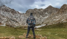 A person wearing a backpack stands on a grassy landscape, looking at snow-dappled mountain peaks under an overcast sky, conveying a sense of adventure and exploration.