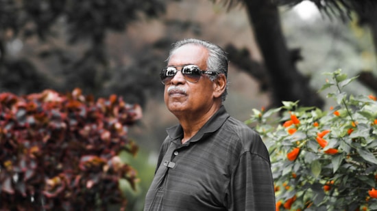 An older man wearing sunglasses, dressed in a dark polo shirt, standing outdoors. He is surrounded by vibrant greenery and colorful flowers, with lush foliage in the background.