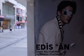 A poster depicting a person in a stylish outfit with text at the bottom. The face is obscured by pasted-on images, creating a fragmented look. The background shows the street with various shops and a modern urban setting.