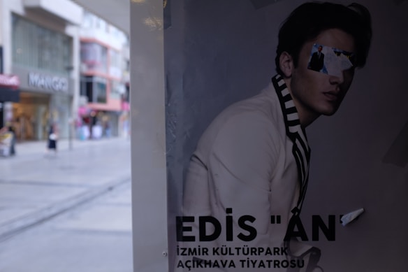 A poster depicting a person in a stylish outfit with text at the bottom. The face is obscured by pasted-on images, creating a fragmented look. The background shows the street with various shops and a modern urban setting.