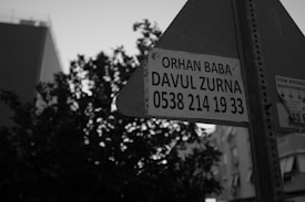 A black and white photograph featuring a street sign with an advertisement for 'Orhan Baba Davul Zurna' including a contact number. The background includes the silhouette of a tree and some buildings.