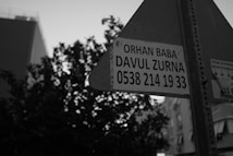 A black and white photograph featuring a street sign with an advertisement for 'Orhan Baba Davul Zurna' including a contact number. The background includes the silhouette of a tree and some buildings.