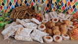 A vibrant array of pastries, cookies, and fritters, dusted with powdered sugar are displayed on a wooden surface. Two glasses of champagne stand in the background, surrounded by colorful party streamers and a harlequin-patterned backdrop, evoking a festive atmosphere.