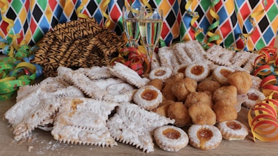 A vibrant array of pastries, cookies, and fritters, dusted with powdered sugar are displayed on a wooden surface. Two glasses of champagne stand in the background, surrounded by colorful party streamers and a harlequin-patterned backdrop, evoking a festive atmosphere.