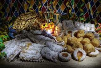 A festive arrangement featuring a variety of pastries dusted with powdered sugar, accompanied by two filled glasses of champagne. The background is decorated with colorful streamers and a harlequin-patterned cloth.