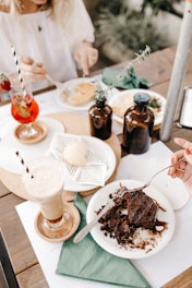 A table set with a variety of desserts and drinks. There are two glasses, one with a red beverage garnished with a strawberry and the other with a frothy latte. Plates hold a piece of chocolate cake with syrup and a pastry with striped napkin. Two brown glass bottles with greenery add a rustic touch.