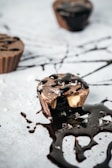 Several chocolate cups are placed on a surface, with one prominently featured partially eaten, revealing a creamy filling inside. Dark chocolate sauce is drizzled artfully across the surface and over the chocolate cups, creating an appetizing display. Salt or sugar crystals are sprinkled around, adding texture to the presentation.