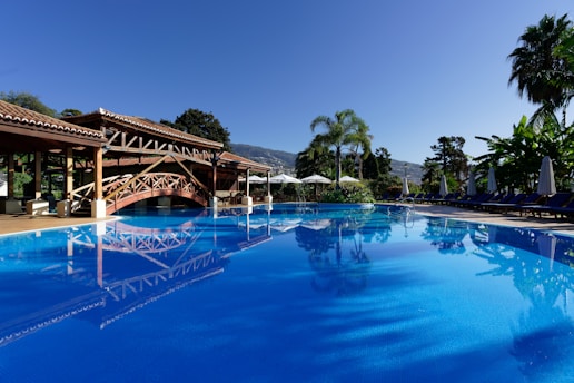 A serene outdoor swimming pool with a calm, reflective water surface. The pool area is surrounded by lush greenery and palm trees, with sun loungers and umbrellas on one side. A charming wooden bridge and a rustic building feature prominently, adding to the tropical resort ambiance under a clear blue sky.