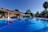 A serene outdoor swimming pool with a calm, reflective water surface. The pool area is surrounded by lush greenery and palm trees, with sun loungers and umbrellas on one side. A charming wooden bridge and a rustic building feature prominently, adding to the tropical resort ambiance under a clear blue sky.