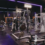 A gym equipped with various weightlifting machines, benches, and barbells. The environment is dimly lit with neon purple lights accentuating the modern gym interior.