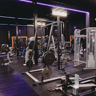 A gym equipped with various weightlifting machines, benches, and barbells. The environment is dimly lit with neon purple lights accentuating the modern gym interior.