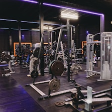 A gym equipped with various weightlifting machines, benches, and barbells. The environment is dimly lit with neon purple lights accentuating the modern gym interior.