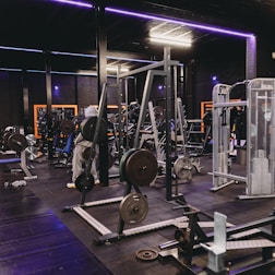 A gym equipped with various weightlifting machines, benches, and barbells. The environment is dimly lit with neon purple lights accentuating the modern gym interior.