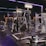 A gym equipped with various weightlifting machines, benches, and barbells. The environment is dimly lit with neon purple lights accentuating the modern gym interior.