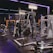 A gym equipped with various weightlifting machines, benches, and barbells. The environment is dimly lit with neon purple lights accentuating the modern gym interior.
