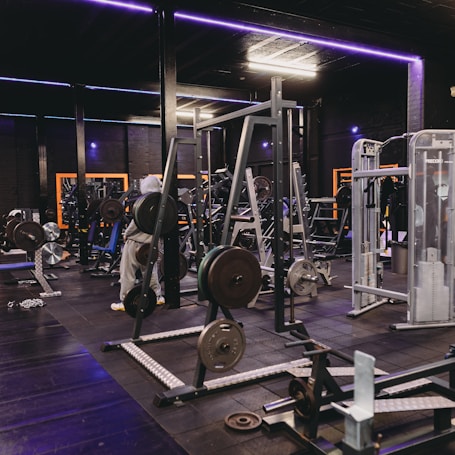 A gym equipped with various weightlifting machines, benches, and barbells. The environment is dimly lit with neon purple lights accentuating the modern gym interior.