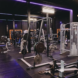 A gym equipped with various weightlifting machines, benches, and barbells. The environment is dimly lit with neon purple lights accentuating the modern gym interior.
