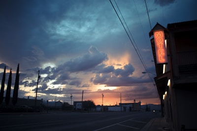 A twilight scene with a dramatic sky filled with dark clouds and a glowing sunset. A roadside motel sign is illuminated in neon red on the right side, casting a warm glow. Tall trees and power lines are silhouetted against the sky, creating a tranquil yet somewhat mysterious atmosphere.