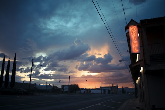 A twilight scene with a dramatic sky filled with dark clouds and a glowing sunset. A roadside motel sign is illuminated in neon red on the right side, casting a warm glow. Tall trees and power lines are silhouetted against the sky, creating a tranquil yet somewhat mysterious atmosphere.