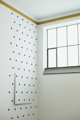 A minimalist interior corner with a white wall dotted with a pattern of small blue squares. The adjacent wall features a grid window with metal framing, and there is a strip of brown tiles at the top edge of the dotted wall. The room has a clean and modern aesthetic.