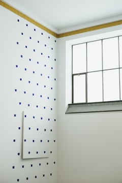 A minimalist interior corner with a white wall dotted with a pattern of small blue squares. The adjacent wall features a grid window with metal framing, and there is a strip of brown tiles at the top edge of the dotted wall. The room has a clean and modern aesthetic.