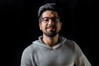 Man with slicked back black hair and black rimmed glasses in front of a black background