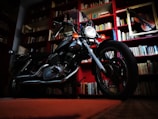 A motorcycle is parked indoors in front of a bookshelf filled with books. The room has red shelves and there is a framed violin on the wall.