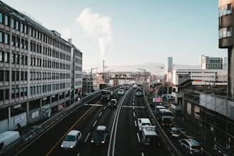 A busy urban road with multiple cars driving in both directions, surrounded by tall buildings. A plume of smoke rises from a chimney in the background, and the sky is clear with a subtle gradient from light to darker blue near the horizon. Traffic signs and streetlights are visible along the road, adding to the organized cityscape.