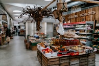A rustic indoor market scene featuring various types of fresh produce displayed on wooden crates. The setting includes fruits, vegetables, and bottled products. A unique decorative tree structure hangs overhead, adding to the artisanal atmosphere. On the left, shelves contain packaged goods, and a person is seen holding a shopping bag.