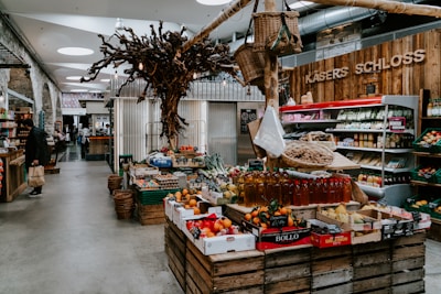 A rustic indoor market scene featuring various types of fresh produce displayed on wooden crates. The setting includes fruits, vegetables, and bottled products. A unique decorative tree structure hangs overhead, adding to the artisanal atmosphere. On the left, shelves contain packaged goods, and a person is seen holding a shopping bag.