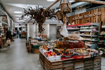 A rustic indoor market scene featuring various types of fresh produce displayed on wooden crates. The setting includes fruits, vegetables, and bottled products. A unique decorative tree structure hangs overhead, adding to the artisanal atmosphere. On the left, shelves contain packaged goods, and a person is seen holding a shopping bag.