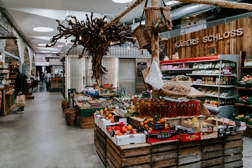 A rustic indoor market scene featuring various types of fresh produce displayed on wooden crates. The setting includes fruits, vegetables, and bottled products. A unique decorative tree structure hangs overhead, adding to the artisanal atmosphere. On the left, shelves contain packaged goods, and a person is seen holding a shopping bag.