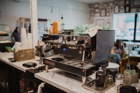 A coffee shop counter features a commercial espresso machine with several green cups stacked on top. Various items are on the counter, such as disposable cups, drink syrups, and utensils. In the background, a person is seated, possibly engaged in conversation or relaxing.