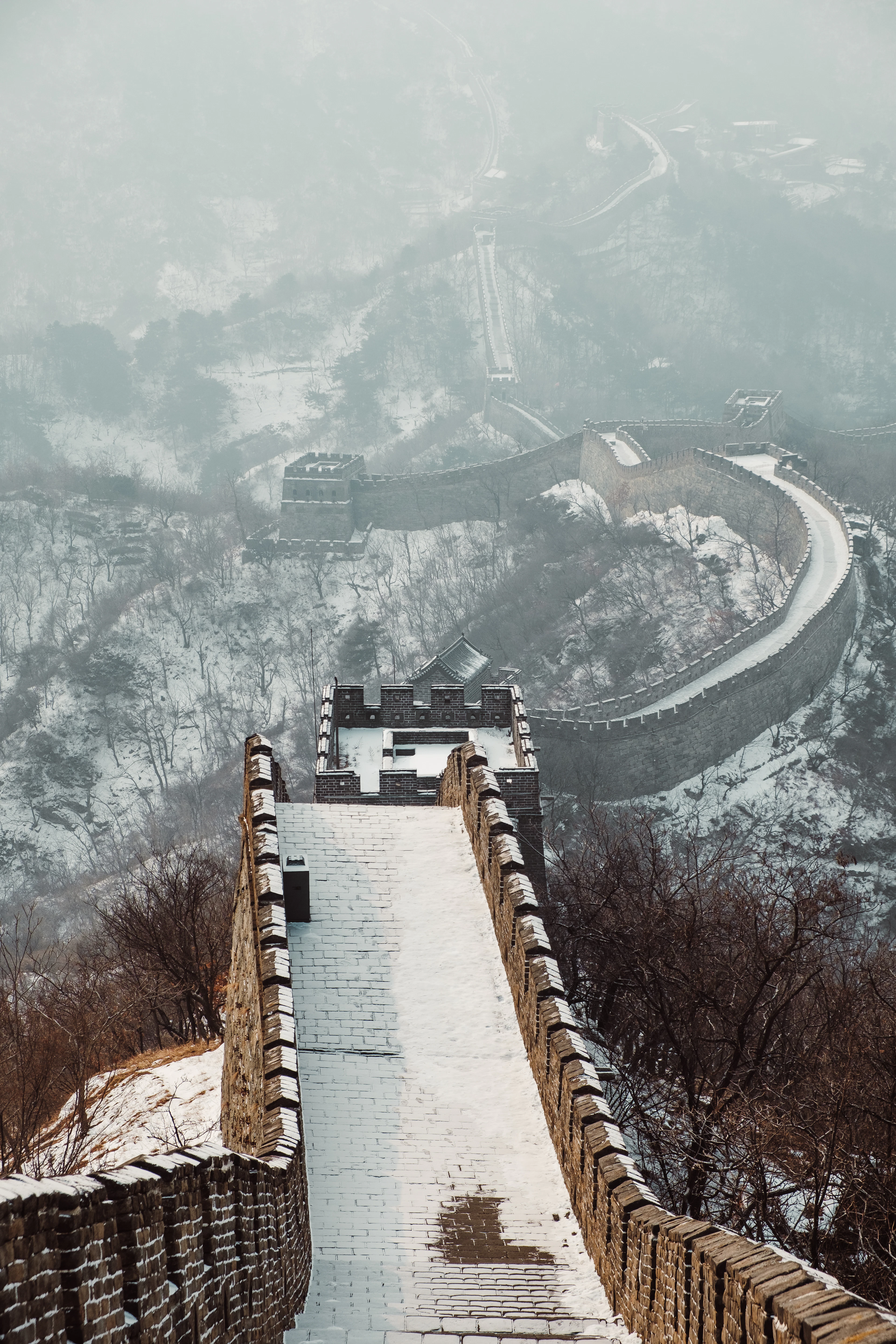 The Great Wall of China under fresh snow, its empty rampart and watchtowers curving away into freezing mist above bare, snow-dusted hillsides.